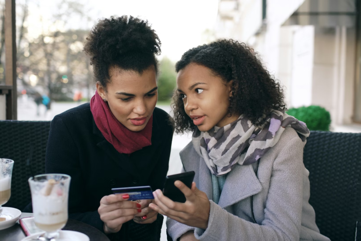 Two women using a smart phone and credit card while seated at a café.