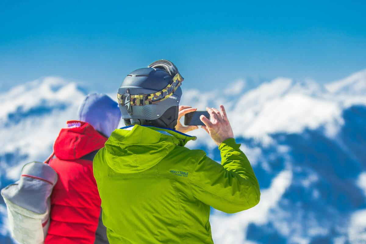 A skier wearing a helmet and winter jacket taking a photo with a smartphone on a snowy mountain.