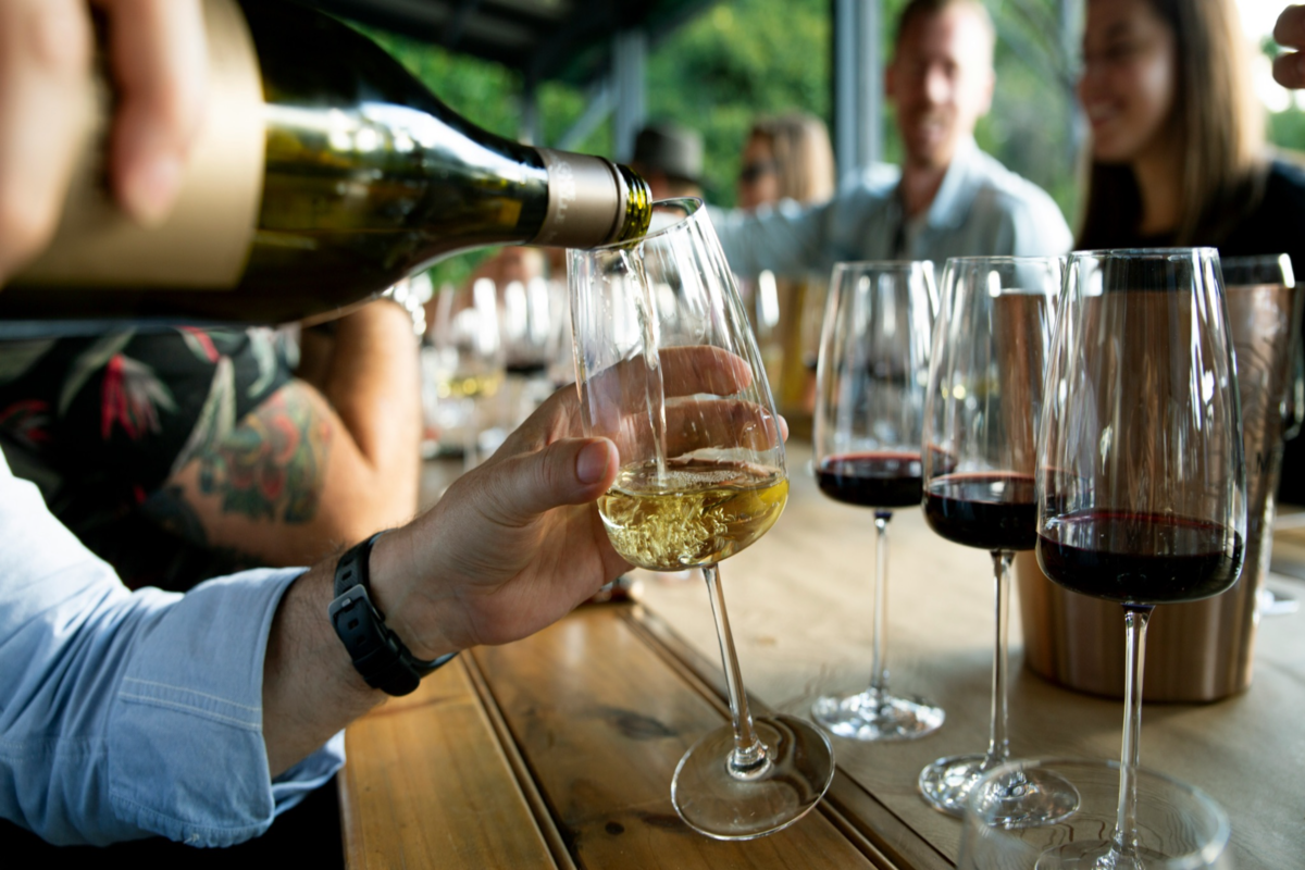 A hand pours white wine from a bottle into a glass, with several glasses of red wine lined up on a wooden table.