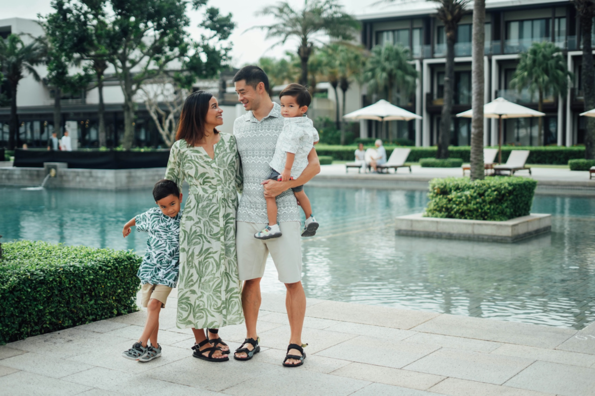 Parents with two young children at a resort with a pool and lounges in the background.