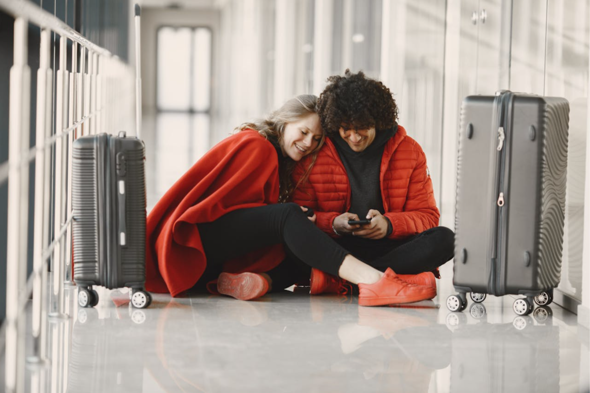 A couple sitting on the floor at the airport, next to suitcases, smiling while looking at a phone. 