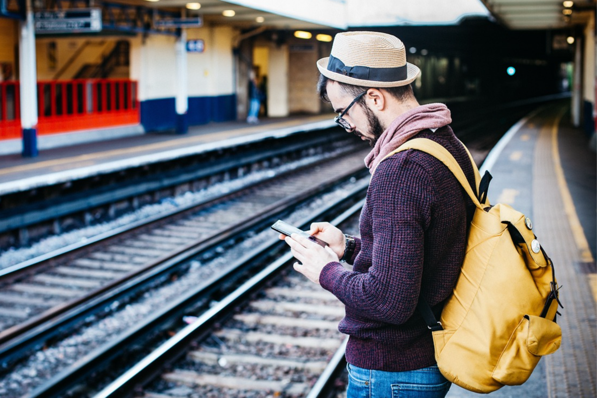 A traveler with a yellow backpack checks their phone while waiting on a train platform.