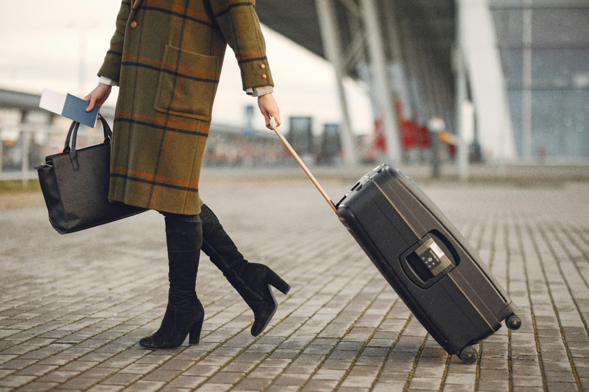 A close-up of a woman from the waist down pulling a suitcase behind her as she walks toward the airport entrance.
