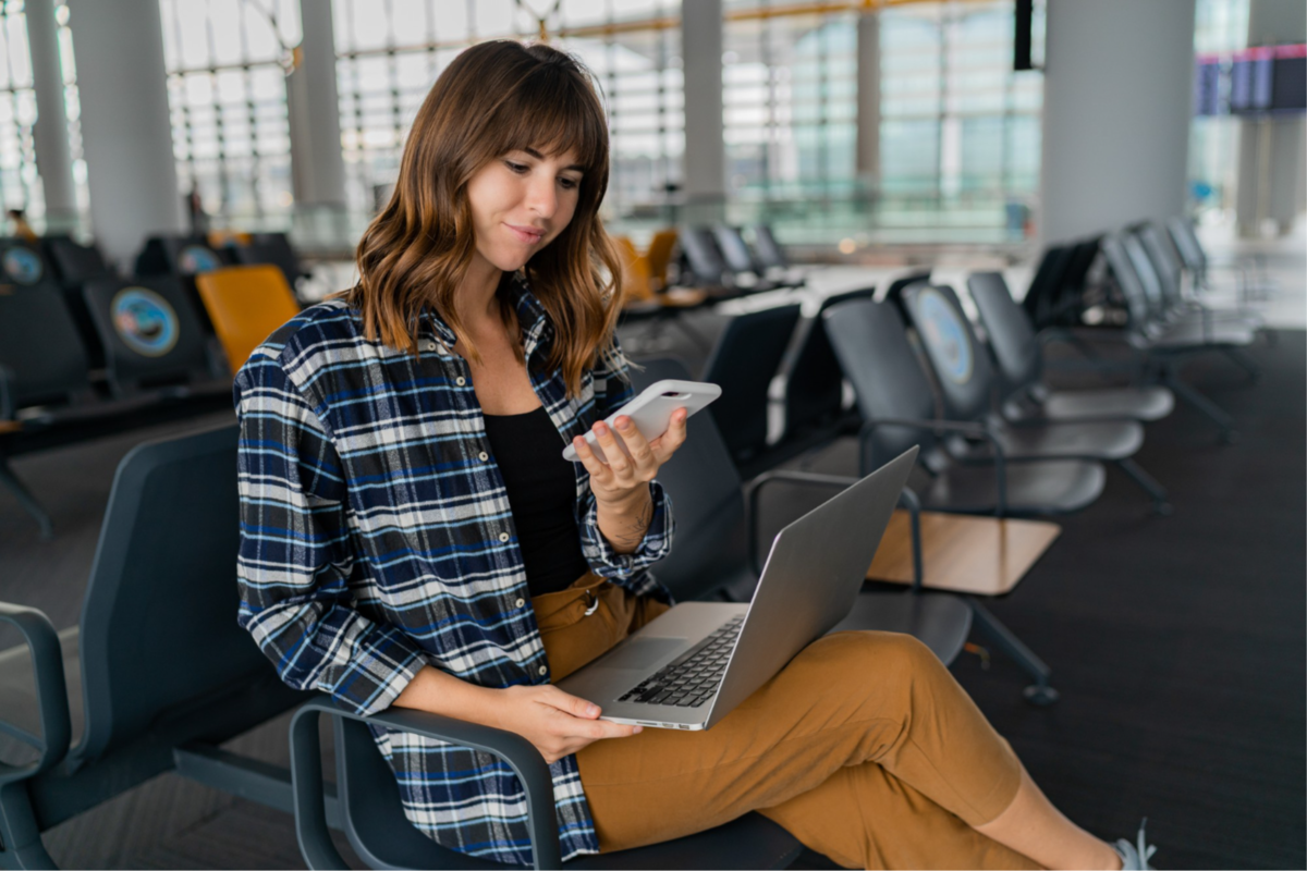 A woman sitting in an empty airport with a laptop in her lap, holding a cell phone in one hand.