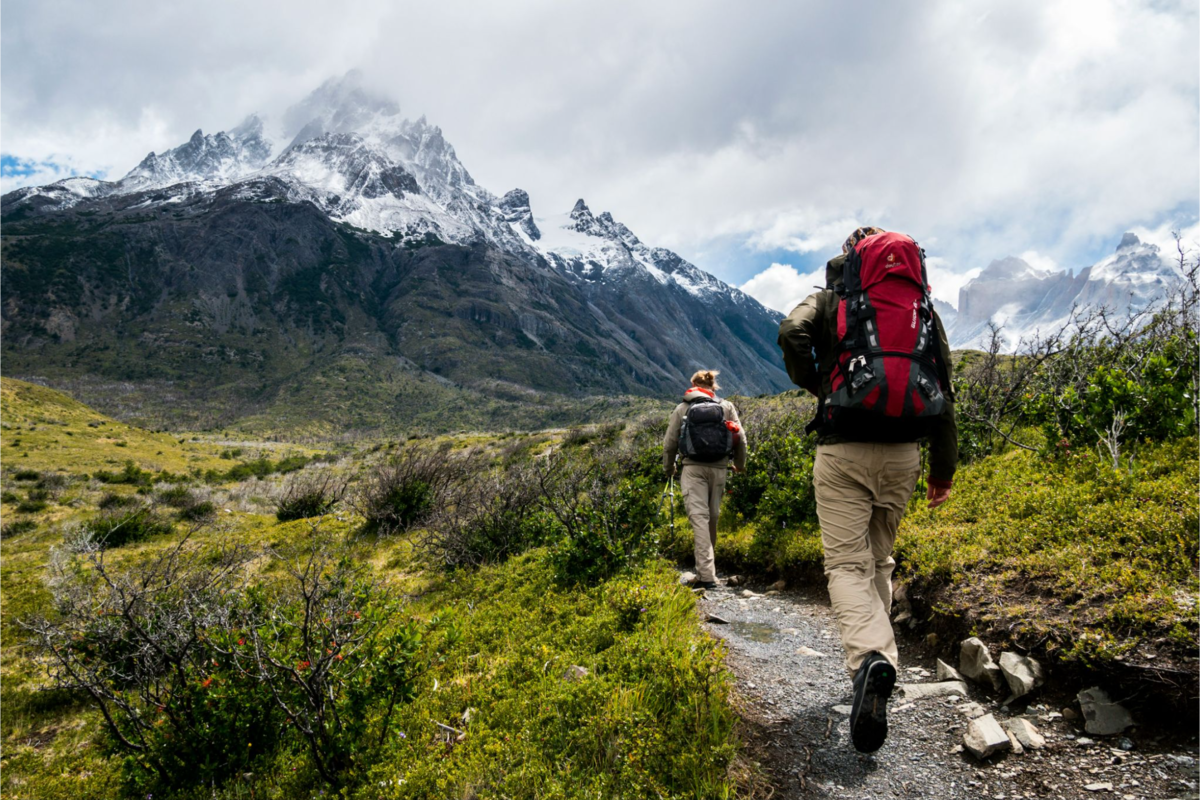 Two hikers trekking toward the towering, snow-capped mountains of Patagonia, surrounded by rugged peaks and open wilderness.