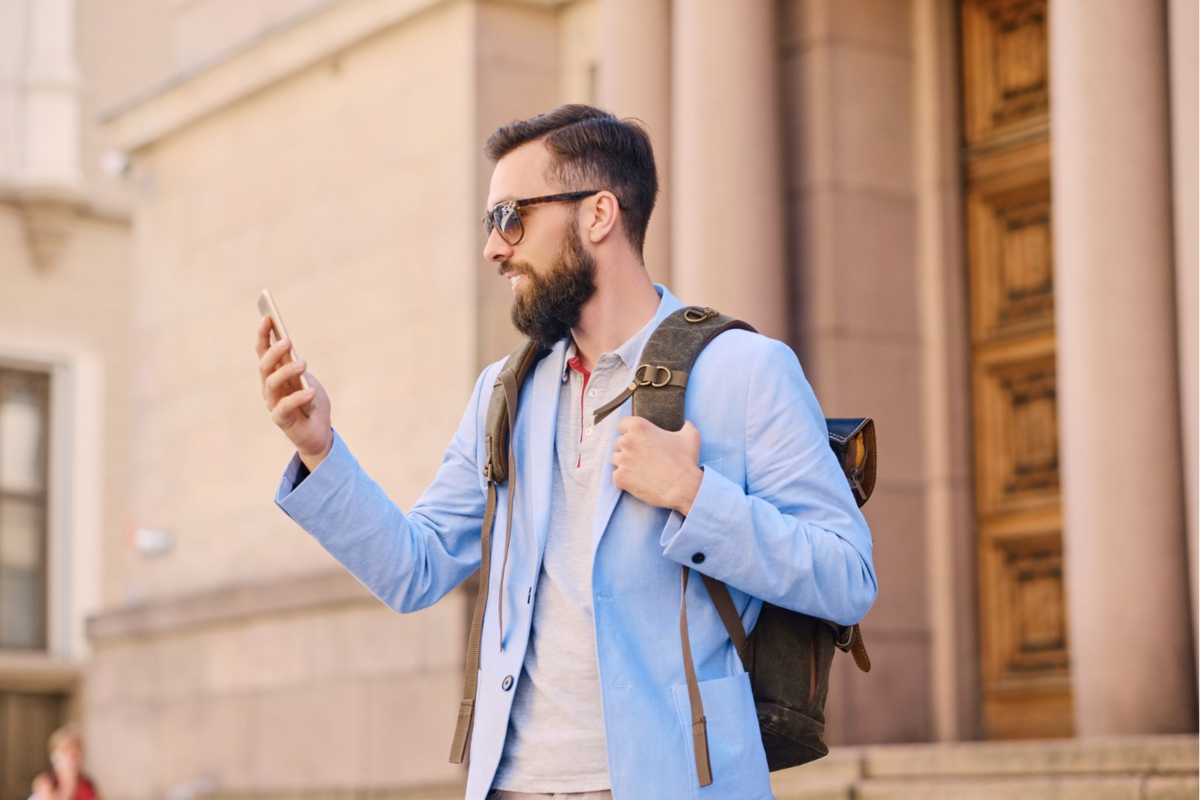 A traveler with a backpack looking at their smartphone while standing in front of a stone building.