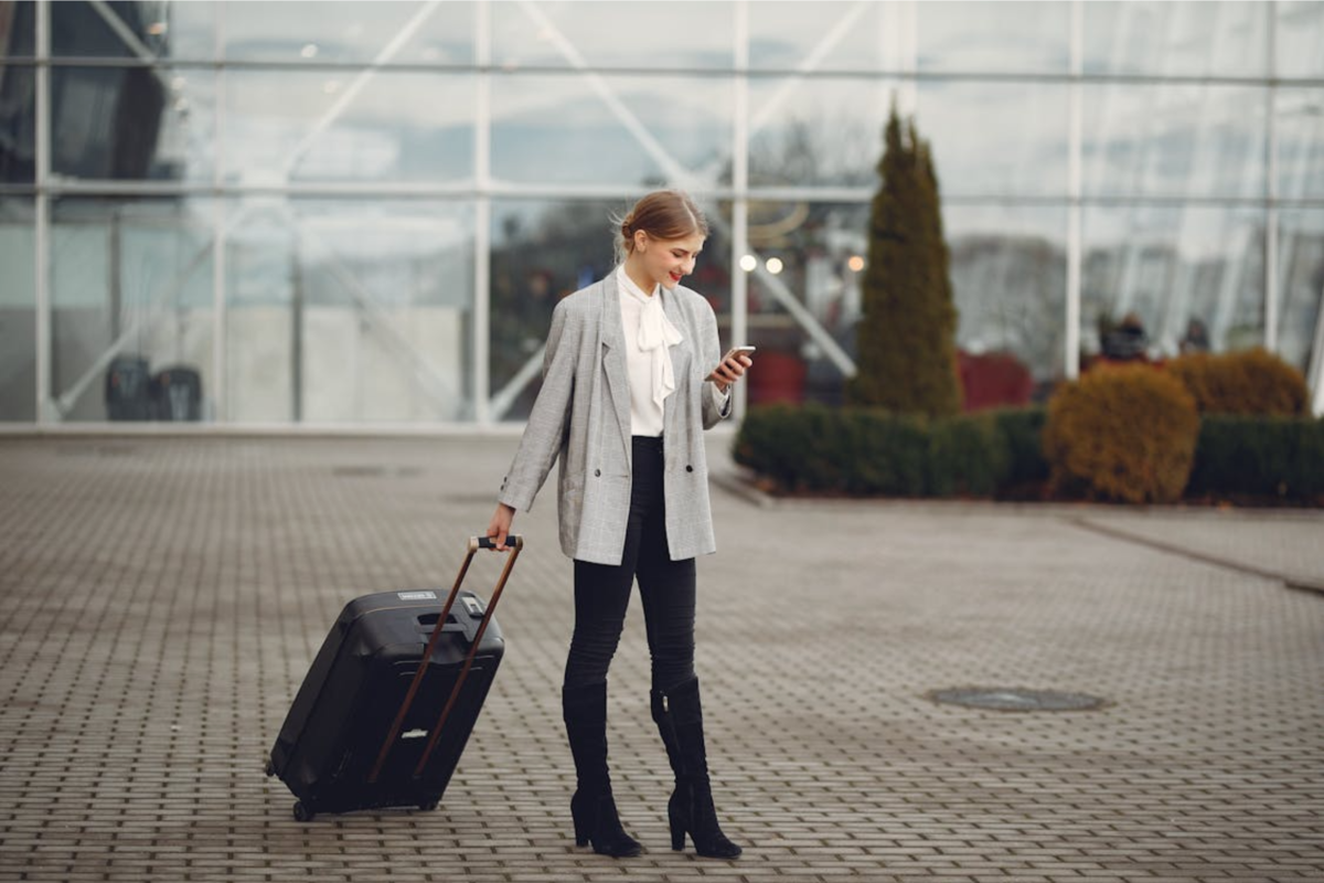 A woman holding the handle of her carry-on suitcase looking down at her phone.